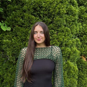 May include: A dark green crochet shrug featuring an open, net-like stitch pattern with long sleeves and a wide, horizontal neckline. The garment is worn over a solid black tank top, showcasing the intricate, textured yarn construction throughout.