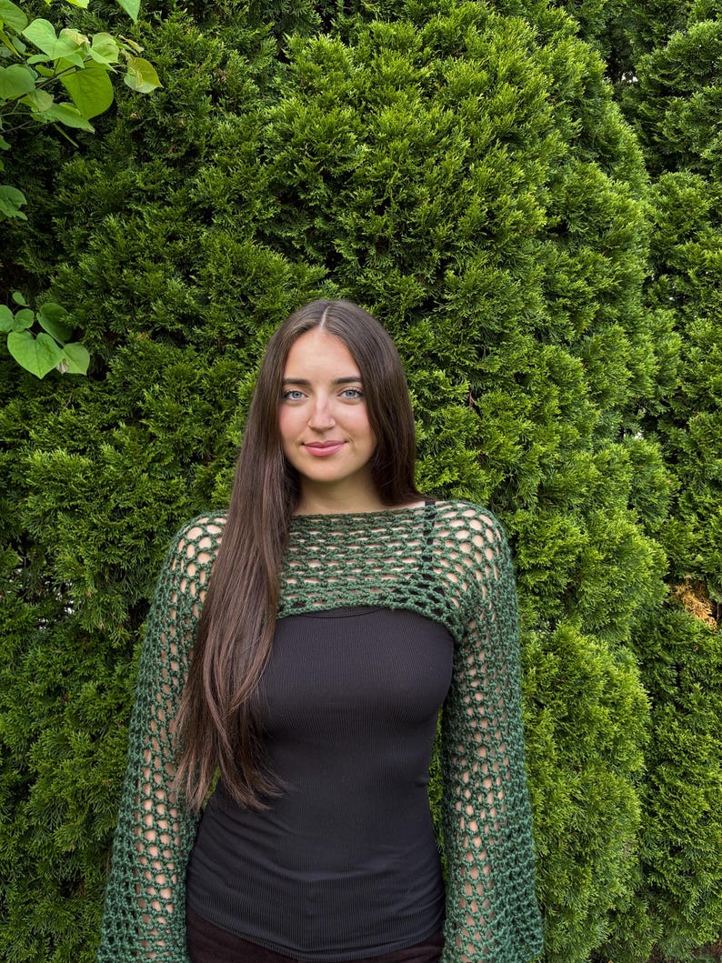 May include: A dark green crochet shrug featuring an open, net-like stitch pattern with long sleeves and a wide, horizontal neckline. The garment is worn over a solid black tank top, showcasing the intricate, textured yarn construction throughout.