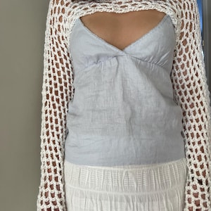 May include: A white long-sleeve crochet shrug with an open net pattern worn over a light blue camisole top and a white textured skirt. The shrug features a wide neckline and long sleeves, while the camisole has a V-neckline and thin straps.