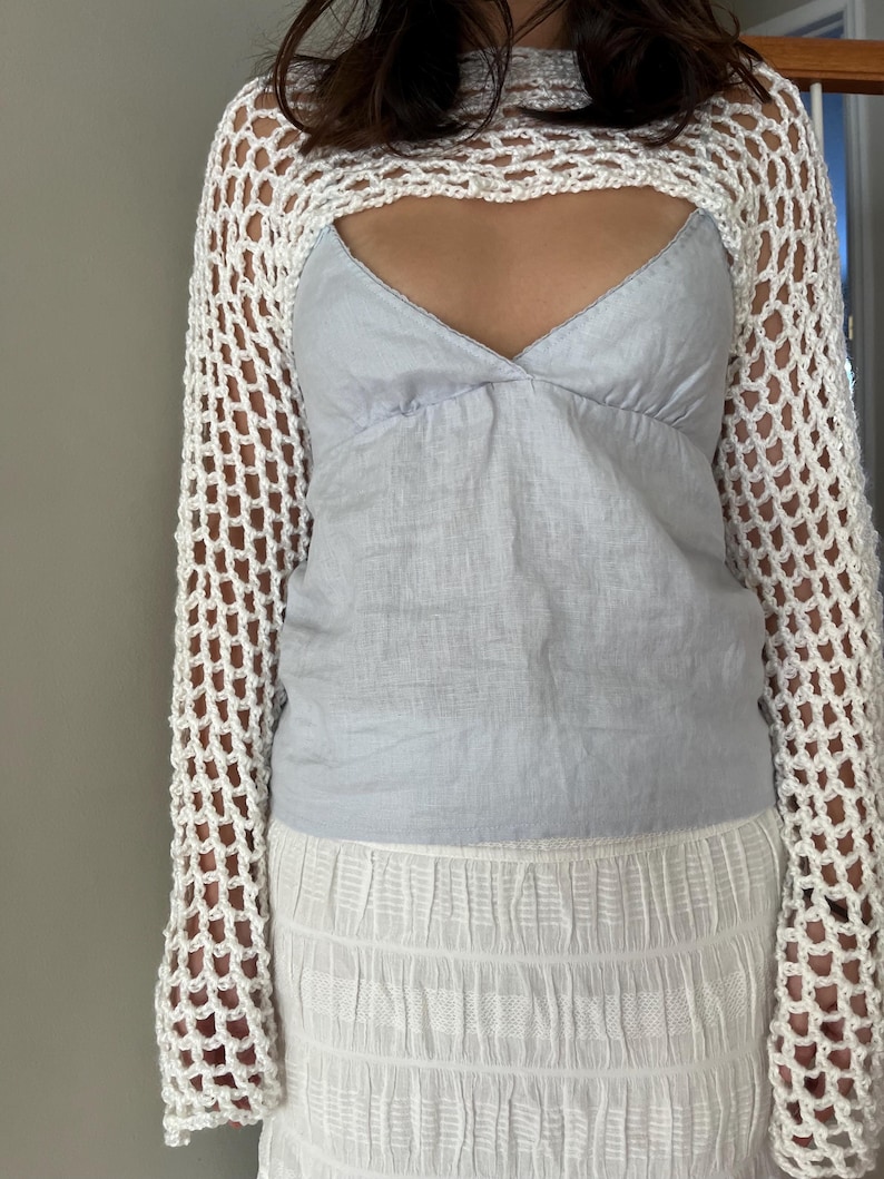 May include: A white long-sleeve crochet shrug with an open net pattern worn over a light blue camisole top and a white textured skirt. The shrug features a wide neckline and long sleeves, while the camisole has a V-neckline and thin straps.