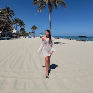 May include: A woman wearing a white, open-weave cover-up dress with long sleeves, walking on a sandy beach. The dress is knee-length and has a loose fit. The background includes palm trees, a clear blue sky, and the ocean.