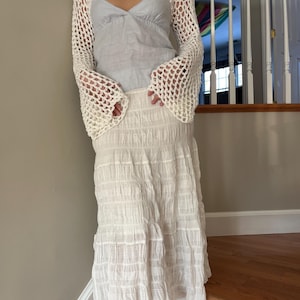 May include: A white long-sleeved crochet shrug with a wide-knit pattern and flared sleeves worn over a light blue sleeveless top with a V-neckline, paired with a long white skirt featuring a textured, crinkled, and horizontally pleated fabric.