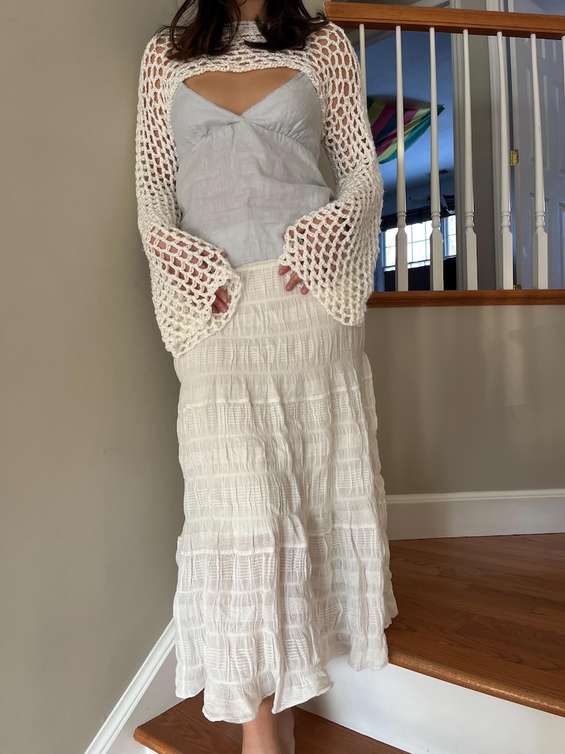 May include: A white long-sleeved crochet shrug with a wide-knit pattern and flared sleeves worn over a light blue sleeveless top with a V-neckline, paired with a long white skirt featuring a textured, crinkled, and horizontally pleated fabric.