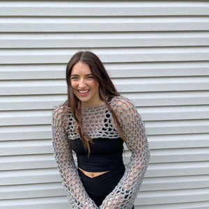 May include: A grey crochet bolero featuring an open-knit mesh pattern with long sleeves and a cropped length, worn over a solid black sleeveless top and black leggings. The garment is constructed from yarn with a visible, textured weave.
