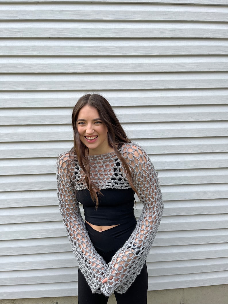 May include: A grey crochet bolero featuring an open-knit mesh pattern with long sleeves and a cropped length, worn over a solid black sleeveless top and black leggings. The garment is constructed from yarn with a visible, textured weave.