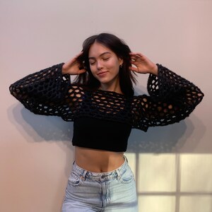 May include: A black long-sleeve crochet shrug featuring an open, loose-knit mesh pattern with wide sleeves, worn over a solid black cropped tank top and light-wash blue denim jeans. The garment has a cropped length and a wide, rounded neckline.