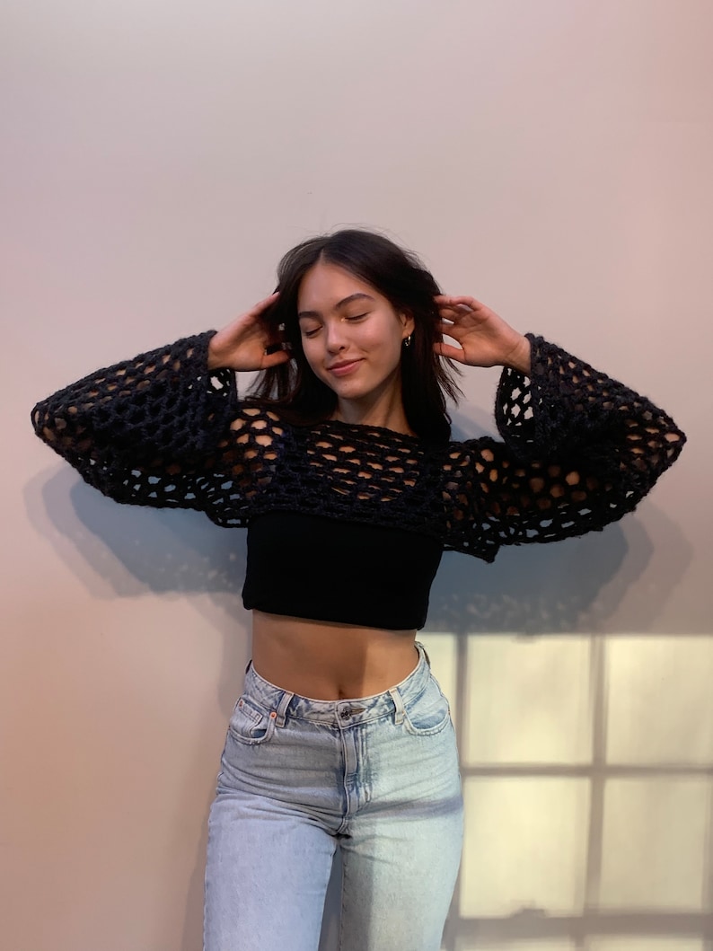 May include: A black long-sleeve crochet shrug featuring an open, loose-knit mesh pattern with wide sleeves, worn over a solid black cropped tank top and light-wash blue denim jeans. The garment has a cropped length and a wide, rounded neckline.