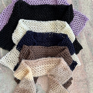 May include: A collection of six crocheted scarves stacked vertically, featuring an open-work mesh pattern. The colors include lavender, black, cream, navy blue, dark brown, and a light beige, each crafted with a soft, textured yarn finish.
