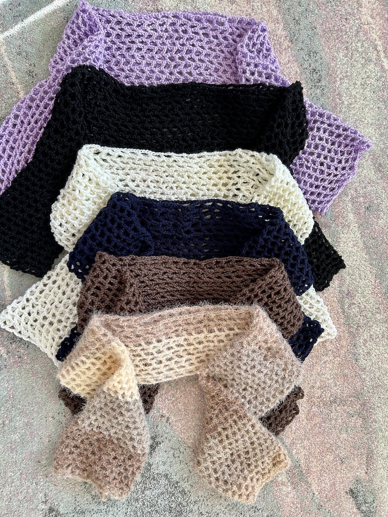 May include: A collection of six crocheted scarves stacked vertically, featuring an open-work mesh pattern. The colors include lavender, black, cream, navy blue, dark brown, and a light beige, each crafted with a soft, textured yarn finish.