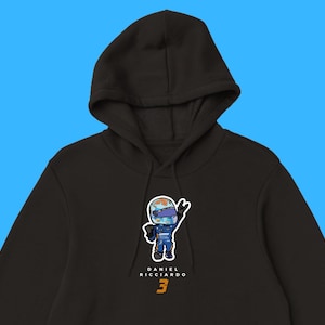 Op de afbeelding: Zwarte hoodie met een cartoonillustratie van Daniel Ricciardo die een racehelm en een racepak draagt. De tekst "DANIEL RICCIARDO 3" is onder de illustratie gedrukt.