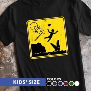 Könnte beinhalten: Schwarzes T-Shirt mit gelbem Warnschild-Grafik, das einen Radfahrer zeigt, der mit einem Krokodil ins Wasser fällt. Der Text "KIDS' SIZE" befindet sich unten, zusammen mit einer Farbauswahl. Das Shirt besteht aus einem weichen Material.
