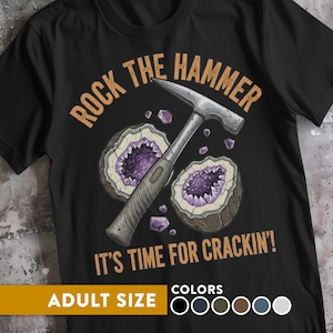 Puede incluir: Camiseta negra con un gráfico que presenta un martillo de geólogo, geodas y el texto "ROCK THE HAMMER IT'S TIME FOR CRACKIN'!" Disponible en tallas de adulto y varios colores.