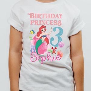May include: White t-shirt featuring a colorful graphic of a mermaid with red hair and a green tail. The shirt has the text "Birthday Princess 3 Sophie" in pink and blue. The shirt is worn by a young child.