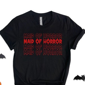 May include: Black t-shirt with the text "Maid of Horror" repeated four times in red. The text is in a bold, stylized font. The shirt is laid flat on a white background with small orange pumpkins and black bats scattered around it.