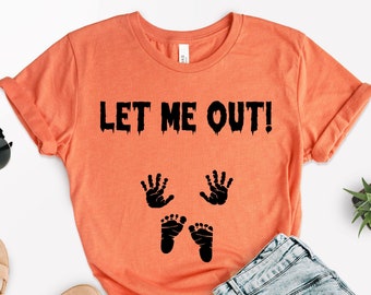 Könnte beinhalten: Orangefarbenes T-Shirt mit dem schwarzen Text "LET ME OUT!" sowie schwarzen Hand- und Fußabdrücken. Das Shirt ist gefaltet, mit Sonnenbrille, Sandalen und Jeans in der Nähe. Eine kleine Pflanze ist ebenfalls sichtbar.