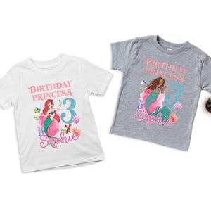 May include: Two short-sleeved t-shirts, one white and one gray, featuring a mermaid graphic. The white shirt has a red-haired mermaid design with the text "Birthday Princess 3 Sophie." The gray shirt has a brown-haired mermaid design with the same text.