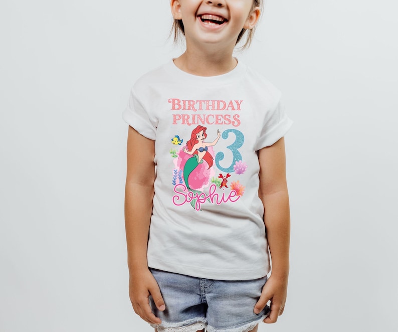 May include: White t-shirt featuring a colorful graphic of a mermaid with red hair and a green tail. The shirt has the text "Birthday Princess 3 Sophie" in pink and blue. The shirt is worn by a young child.