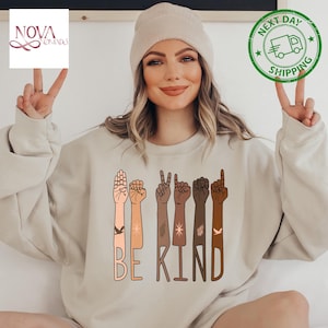 May include: A beige sweatshirt with the words "BE KIND" printed on the front. The words are surrounded by illustrations of hands in different skin tones, each making a different sign language gesture.