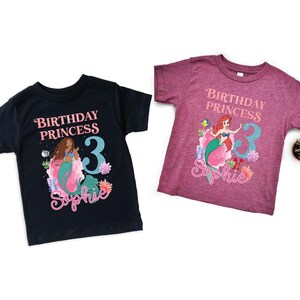 May include: Two short-sleeved t-shirts featuring mermaid graphics. One is navy blue, the other is maroon. Both shirts have the text "Birthday Princess" and a number 3, with a mermaid illustration and a name.