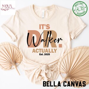 May include: A cream-colored Bella Canvas t-shirt with the text "It's Dr. Walker Actually Est. 2025" in brown and black. The shirt is styled with a knot at the bottom and is surrounded by decorative fans.