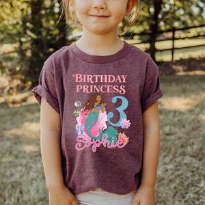 May include: A maroon t-shirt featuring a mermaid graphic with the text "Birthday Princess 3 Sophie". The mermaid has long brown hair and a teal tail. The number 3 is in blue.