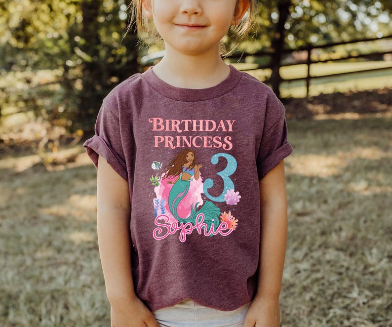 May include: A maroon t-shirt featuring a mermaid graphic with the text "Birthday Princess 3 Sophie". The mermaid has long brown hair and a teal tail. The number 3 is in blue.