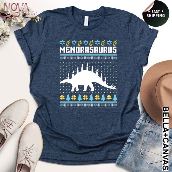 May include: Navy blue t-shirt with a white "MENORASAURUS" design. The design features a dinosaur silhouette with a menorah on its back, surrounded by festive patterns. A fun and unique holiday-themed graphic tee.