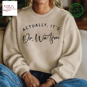 May include: A beige sweatshirt with the text "Actually, it's Dr. Watson" printed in black.