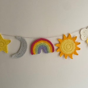 Peut inclure: Une guirlande en crochet avec une étoile jaune, un croissant de lune gris, un arc-en-ciel, un soleil jaune et un nuage blanc avec une goutte de pluie grise.