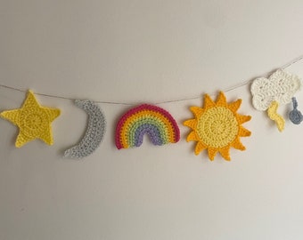 Könnte beinhalten: Eine gehäkelte Girlande mit einem gelben Stern, einem grauen Halbmond, einem Regenbogen, einer gelben Sonne und einer weißen Wolke mit einem grauen Regentropfen.