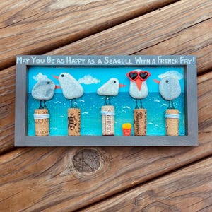 May include: A rectangular wall art piece featuring five pebble seagulls perched on wine corks against a painted blue ocean background. The text reads, "May You Be As Happy As A Seagull With A French Fry!" One seagull wears red heart-shaped glasses.