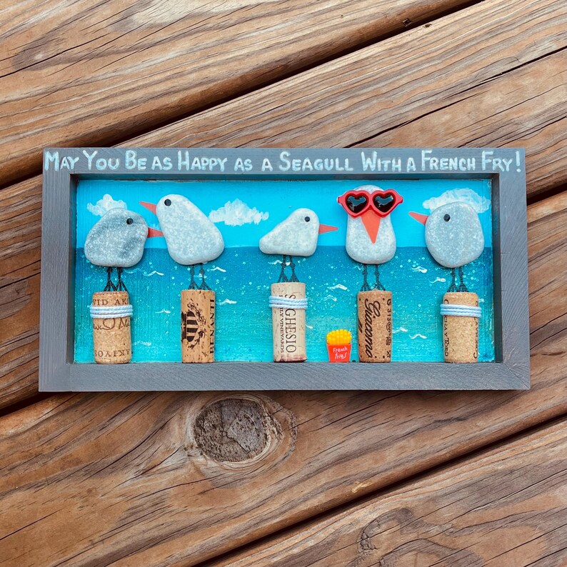 May include: A rectangular wall art piece featuring five pebble seagulls perched on wine corks against a painted blue ocean background. The text reads, "May You Be As Happy As A Seagull With A French Fry!" One seagull wears red heart-shaped glasses.