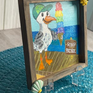 May include: A framed painting of a seagull holding a colorful ice cream cone. The seagull wears a green cap and stands on a pier with the words "SCOOP PATROL" on a sign. The frame is brown, and the background depicts the ocean.
