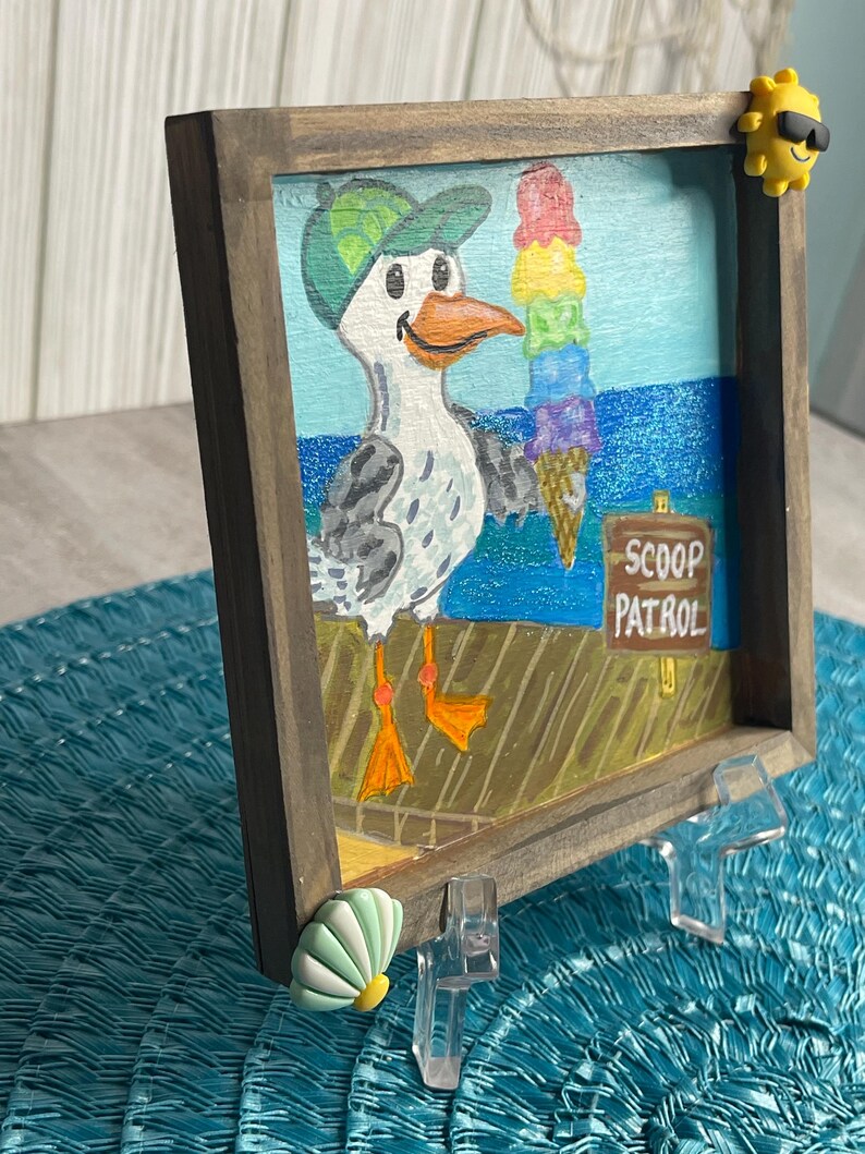 May include: A framed painting of a seagull holding a colorful ice cream cone. The seagull wears a green cap and stands on a pier with the words "SCOOP PATROL" on a sign. The frame is brown, and the background depicts the ocean.