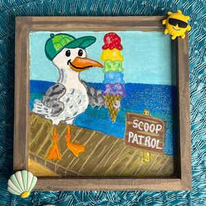 May include: A framed painting of a seagull wearing a green cap, holding a colorful ice cream cone. The painting has a wooden frame and the words "SCOOP PATROL" are visible on a sign. A yellow sun with sunglasses and a seashell are attached to the frame.