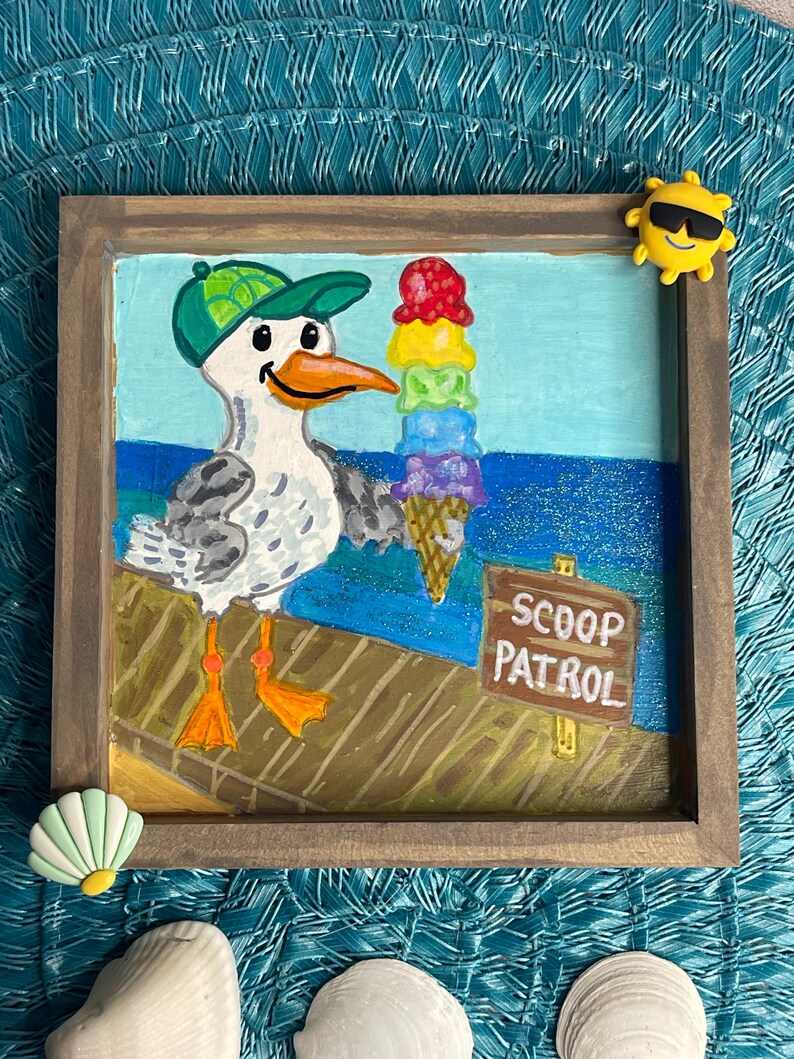 May include: A framed painting of a seagull wearing a green cap, holding a colorful ice cream cone. The painting has a wooden frame and the words "SCOOP PATROL" are visible on a sign. A yellow sun with sunglasses and a seashell are attached to the frame.