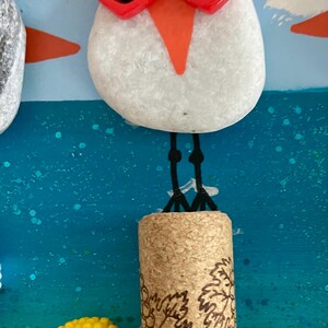 May include: A white stone bird figure wearing red heart-shaped sunglasses with black lenses, standing on a cylindrical cork with a grape vine design. Next to the cork is a small red container filled with yellow french fries on a blue background.