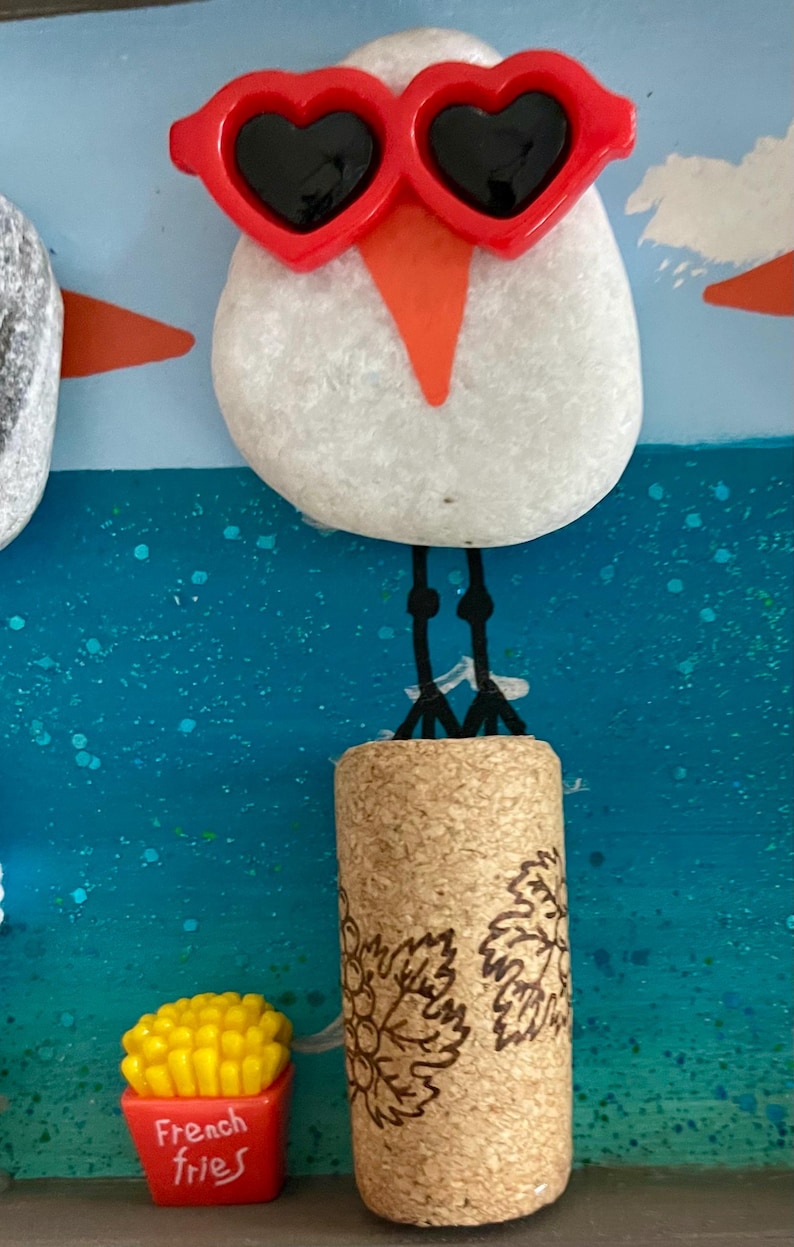 May include: A white stone bird figure wearing red heart-shaped sunglasses with black lenses, standing on a cylindrical cork with a grape vine design. Next to the cork is a small red container filled with yellow french fries on a blue background.