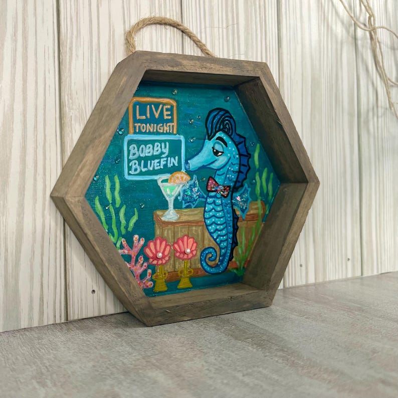 May include: A hexagonal wooden wall art piece featuring a blue seahorse bartender holding a cocktail. The artwork includes the text "LIVE TONIGHT" and "BOBBY BLUEFIN" on a sign. The frame has a rope hanger.