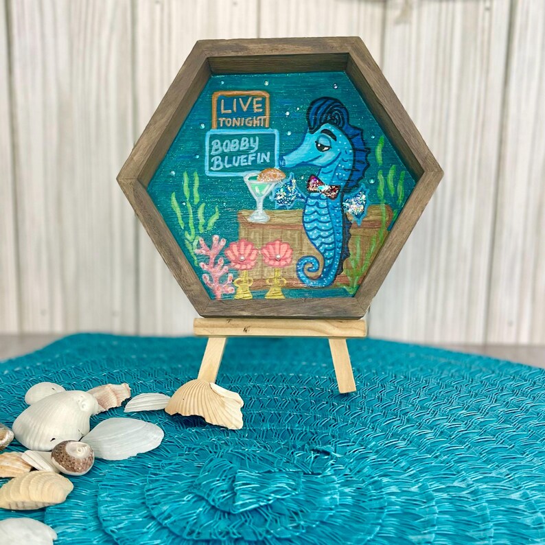 May include: A whimsical, hexagonal wooden art piece featuring a blue seahorse bartender holding a cocktail. The artwork includes the text "LIVE TONIGHT" and "Bobby Bluefin". The seahorse is set against a teal background with coral and seaweed accents. The piece is displayed on a small wooden easel.