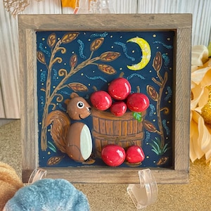 May include: A framed artwork featuring a whimsical scene with a squirrel, apples, and a crescent moon against a dark blue background. The squirrel is brown and white, and the apples are red. The frame is made of wood.
