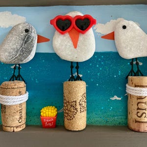 May include: A framed wall art piece featuring three pebble seagulls perched on corks against a painted blue ocean background. The center seagull wears red heart-shaped sunglasses. A small red container labeled French fries sits below.