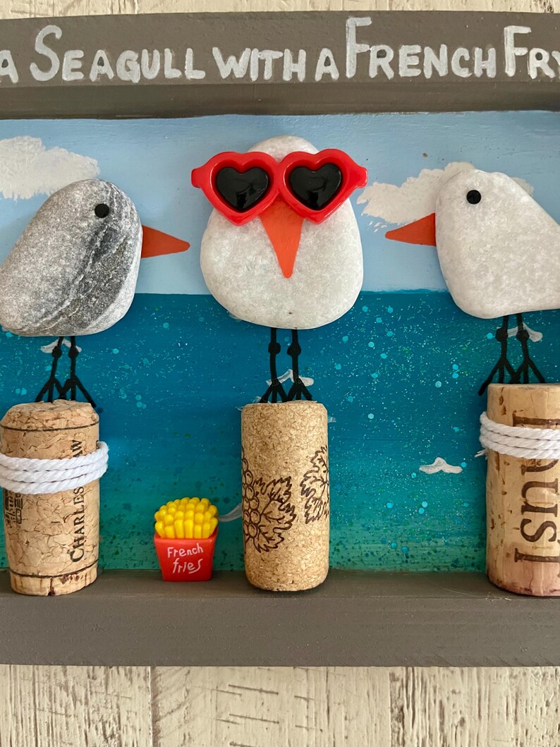 May include: A framed wall art piece featuring three pebble seagulls perched on corks against a painted blue ocean background. The center seagull wears red heart-shaped sunglasses. A small red container labeled French fries sits below.
