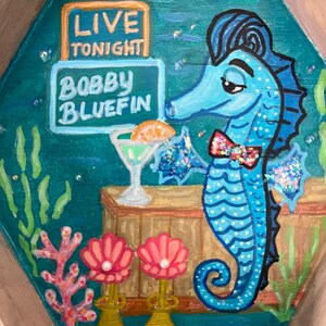 May include: A whimsical, hand-painted wooden tray featuring a blue seahorse with a bow tie, a cocktail, and the text "LIVE TONIGHT BOBBY BLUEFIN." The tray has a teal background with coral and seashell accents, and a wooden frame.