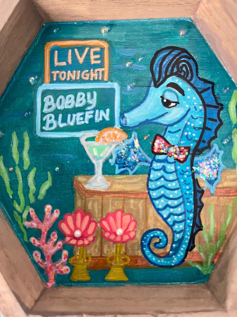 May include: A whimsical, hand-painted wooden tray featuring a blue seahorse with a bow tie, a cocktail, and the text "LIVE TONIGHT BOBBY BLUEFIN." The tray has a teal background with coral and seashell accents, and a wooden frame.