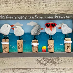 May include: A rectangular wall art piece in a gray frame featuring five pebble seagulls perched on wine corks against a painted blue ocean background. The text reads, May You Be As Happy As A Seagull With A French Fry! One seagull wears red heart-shaped sunglasses.