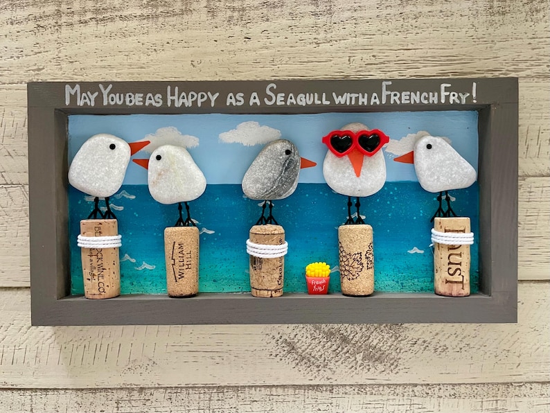 May include: A rectangular wall art piece in a gray frame featuring five pebble seagulls perched on wine corks against a painted blue ocean background. The text reads, May You Be As Happy As A Seagull With A French Fry! One seagull wears red heart-shaped sunglasses.