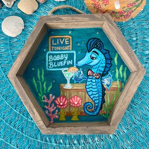 May include: A hexagonal wooden tray with a painted scene of a blue seahorse wearing a bow tie, holding a cocktail. The sign reads "Live Tonight Bobby Bluefin". The background is teal with coral and seaweed.
