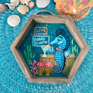 May include: A wooden, hexagonal wall art piece featuring a blue seahorse with a bow tie, holding a cocktail. The sign reads "Live Tonight" and "Bobby Bluefin". The background is a teal ocean scene with coral and seaweed.