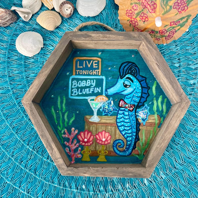 May include: A wooden, hexagonal wall art piece featuring a blue seahorse with a bow tie, holding a cocktail. The sign reads "Live Tonight" and "Bobby Bluefin". The background is a teal ocean scene with coral and seaweed.